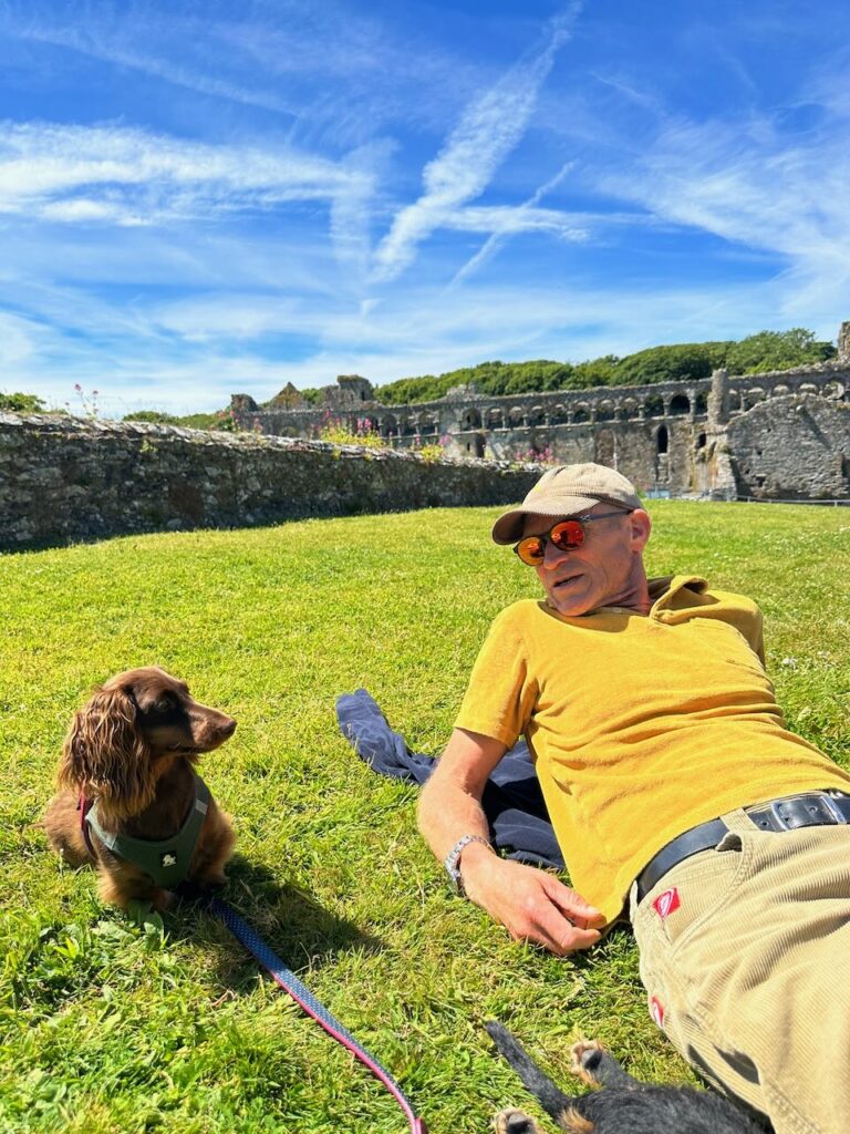 Man in yellow shirt and green cap with dachshund lying on grass with abbey ruins behind them